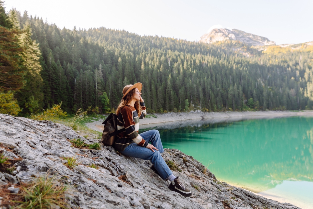 A girl in a sweater, hat and backpack sits on a rock at the edge of a lake with surrounding forest around her.