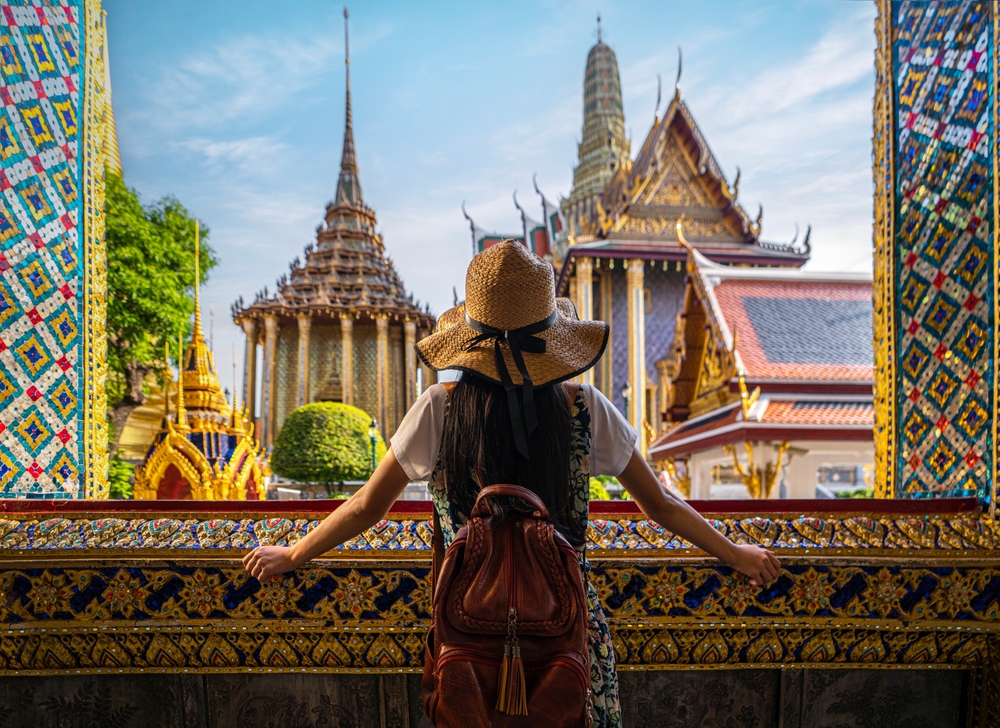 A woman in a straw hat looks out over Asian buildings, framed by ornate designs in a palace open window.