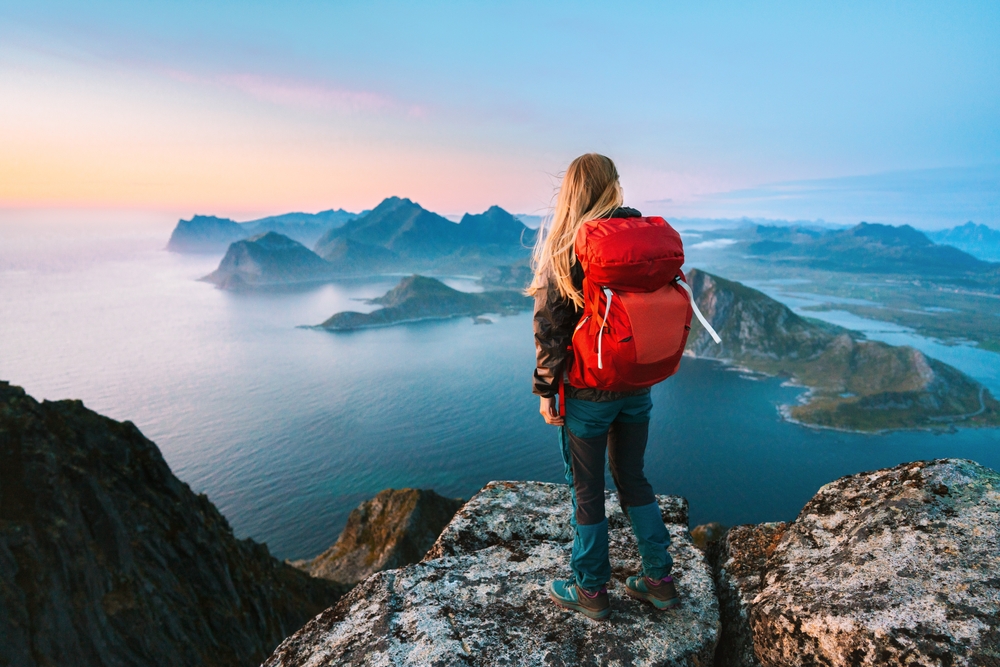 A girl with a red backpack stands on the edge of a cliff and overlooks the mini island formations on the water below.