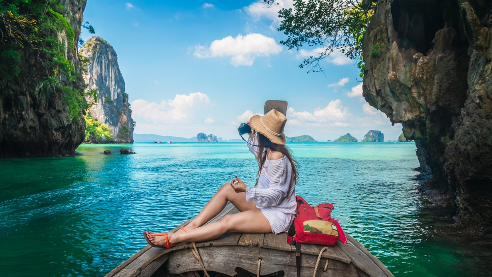A girl in a bathing suit cover up and straw hat looks over the edge of a boat at blue waters and rugged terrain with her camera.