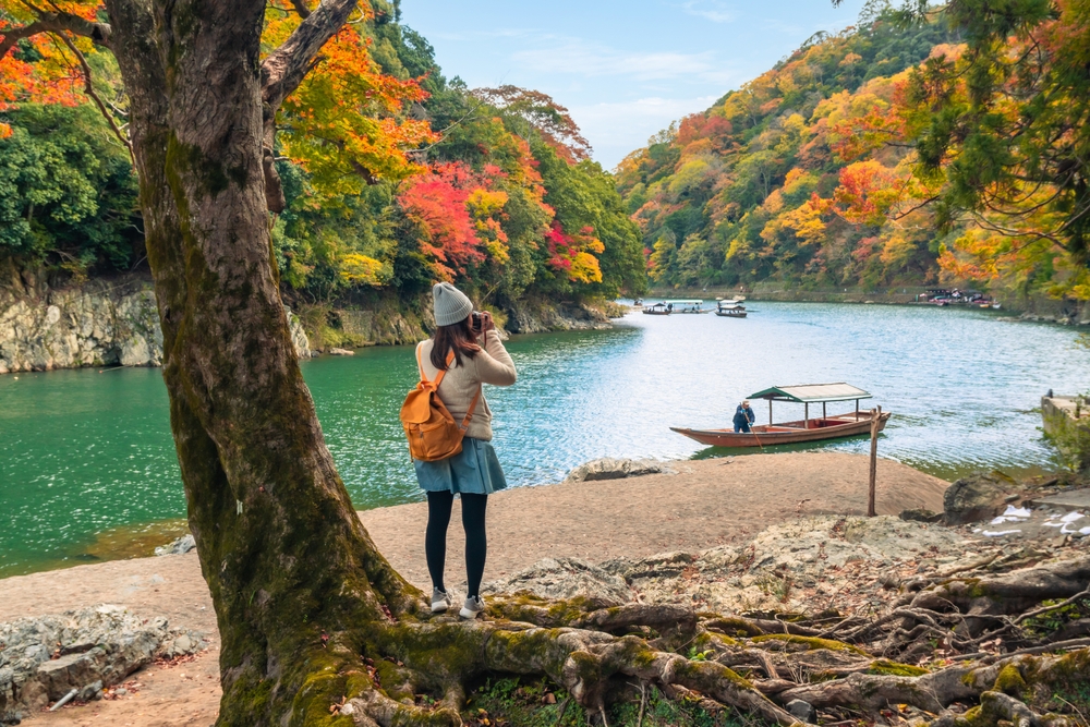 A girl takes a photo of the fall foliage turning orange on the edge of a river while wearing a skirt, tights, an orange backpack, a beanie and a sweater.