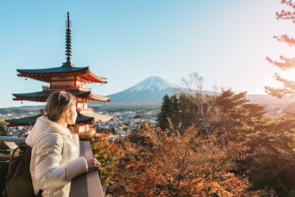 A girl overlooks trees and a temple from a balcony, wearing a larger puffer jacket to stay warm agains the wind chill.