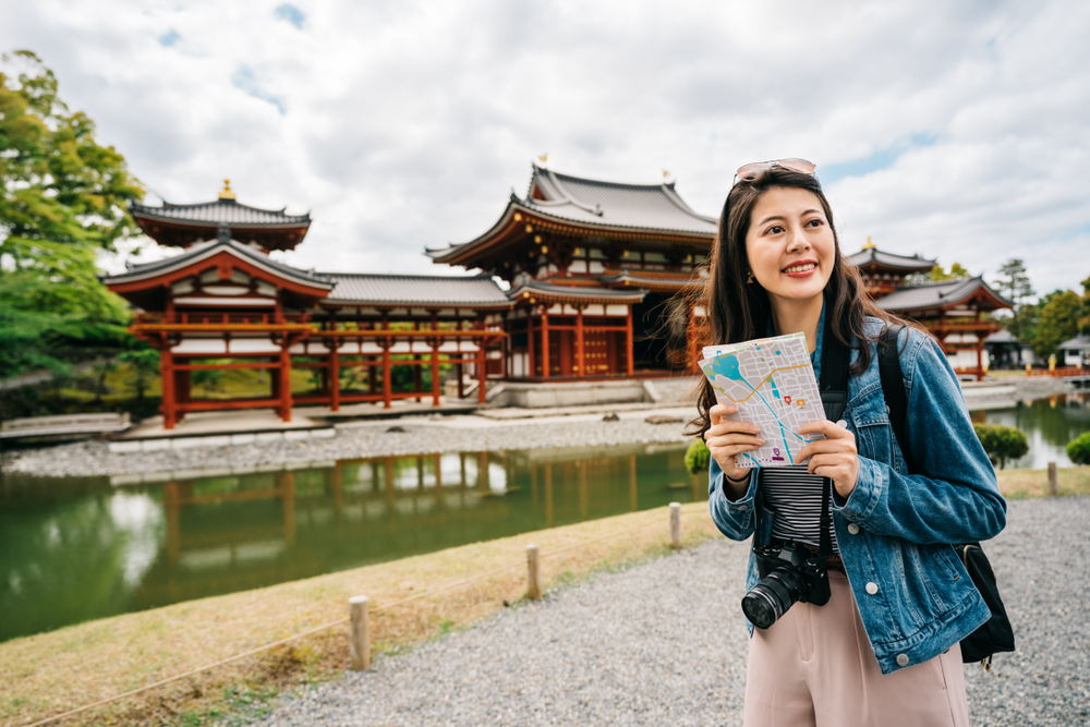 A young girl in a denim jacket and striped shirt shows she knows what to wear to Japan in the fall, as she clutches a map, comfortable, a temple in the background.