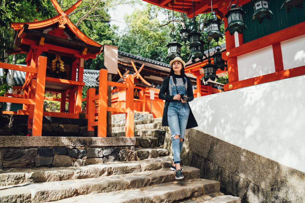 A girl in overalls and a striped shirt with a camera around her neck stands in a temple with Japan, looking over the stone stairs.