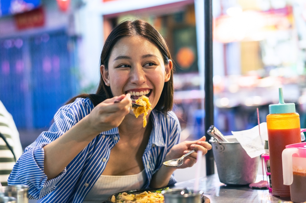 A girl spoons a bite of food into her mouth and laughs while wearing a blue button up and a white cami: she knew what to wear to Japan in the fall in september, as this will keep her cool in the humidity.