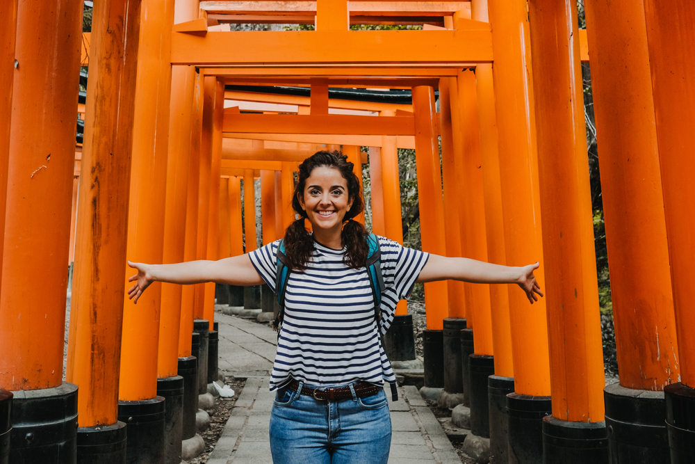 A young girl in a striped shirt and jeans spreads her arms in the orange walkway of a temple and bridge in Japan.