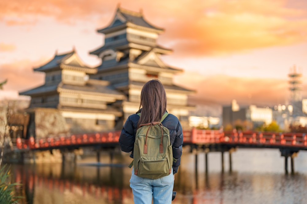 A girl in jeans and a dark blue puffer jacket knows what to wear to Japan in the fall as the sun sets: she is comfortable with her layers and green backpack as she faces a red bridge and cityscape beyond.