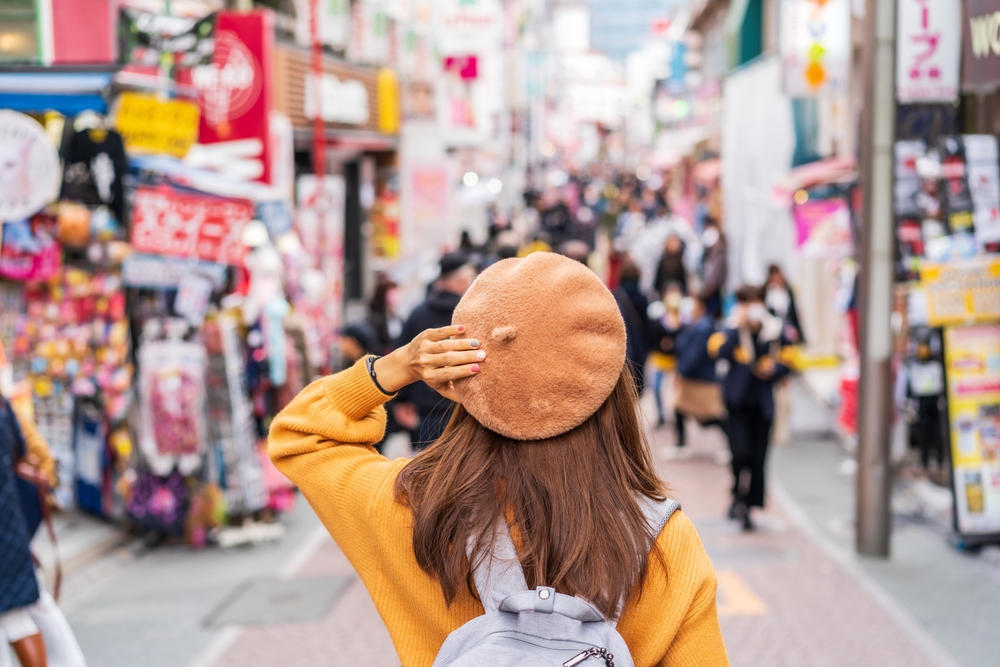 A girl with brown hair reaches up and secures her wool fedora as she wears a backpack, yellow sweater and looks amongst a market.