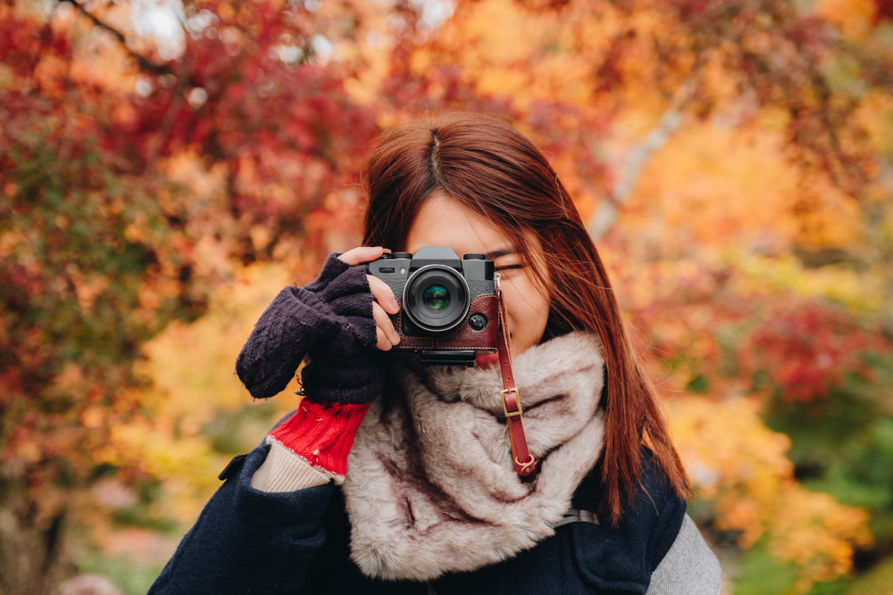 A girl holds her camera up to face us as she wears a thicker scarf, layered hoodies and fingerless gloves: she knows what to wear to Japan in the fall, as it is cooler in November as the fall foliage turns orange behind her.