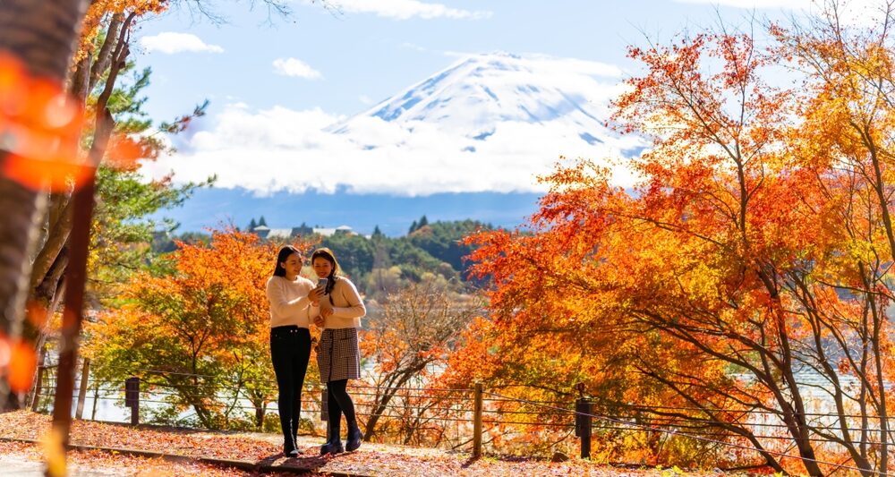 Two girls hover in the fall foliage of Japan, mountains in the background, and they are comfortable because they knew what to wear to Japan in the fall with their long pants, tights, midi skirts and sweaters.