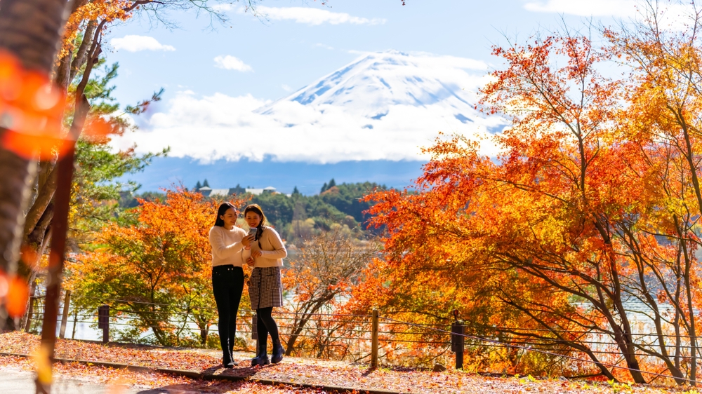 Two girls knew what to wear to Japan in the fall in their boots, tights, midi skirt, and sweaters as they look at their phone amongst snow capped mountains and fall foliage.
