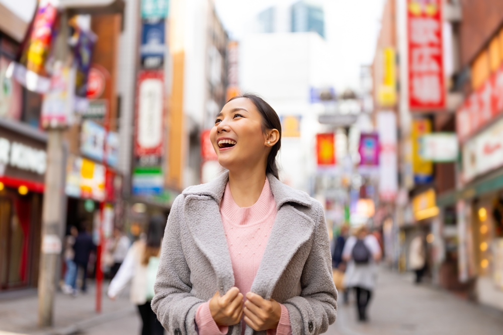 A girl looks up at all the lights of a city, smiling, knowing what to wear to Japan in the fall in her wool overcoat and pink knit sweater.