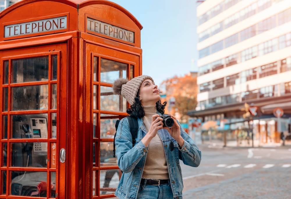 A tourist leans against a red telephone both in a turtle neck and a jean jacket while holding a camera and wearing a beanie.
