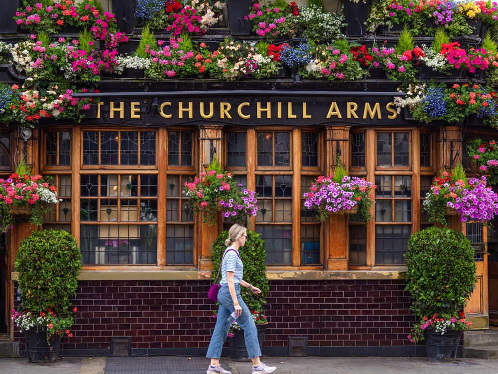 A girl who is walking across the Churchill Arms bar knows what to wear in London in September, as she prepared her layers with jeans, a striped shirt, a jacket that she has off and a crossbody bag.