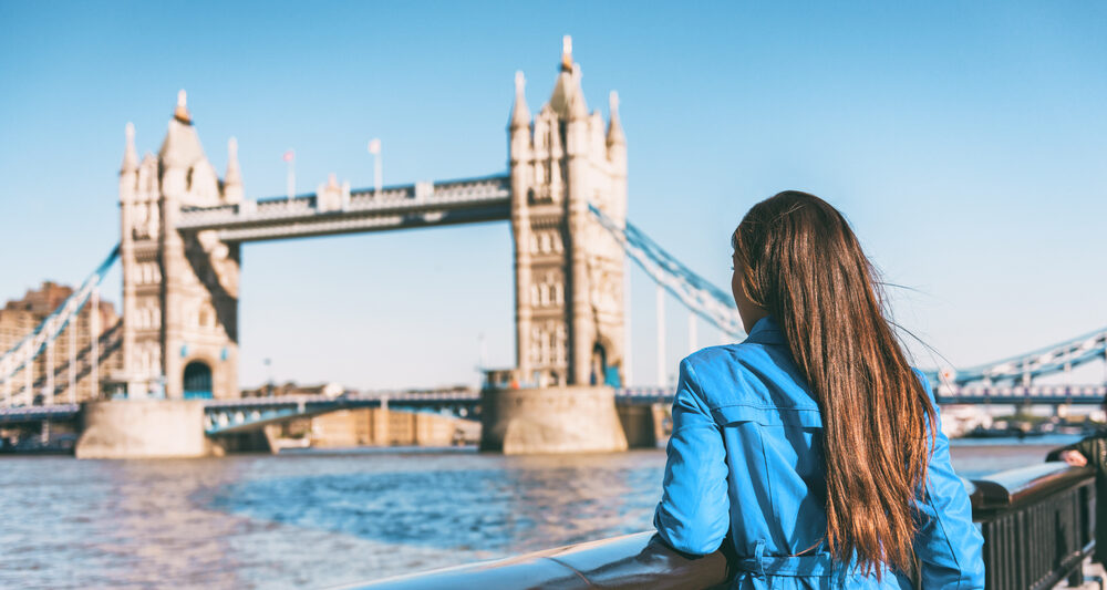 A woman in a blue jacket knows what to wear in London in September as she leans agains the wall on the river Thames facing tower bridge, prepared for the wind off the water.