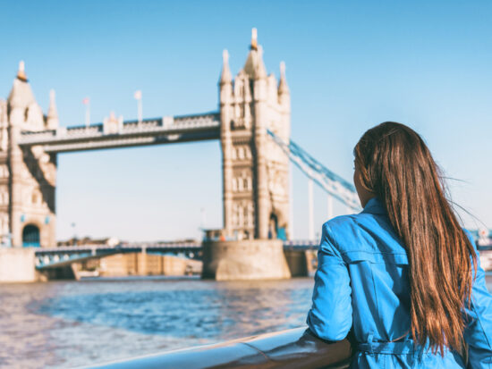 A woman in a blue jacket knows what to wear in London in September as she leans agains the wall on the river Thames facing tower bridge, prepared for the wind off the water.