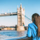A woman in a blue jacket knows what to wear in London in September as she leans agains the wall on the river Thames facing tower bridge, prepared for the wind off the water.