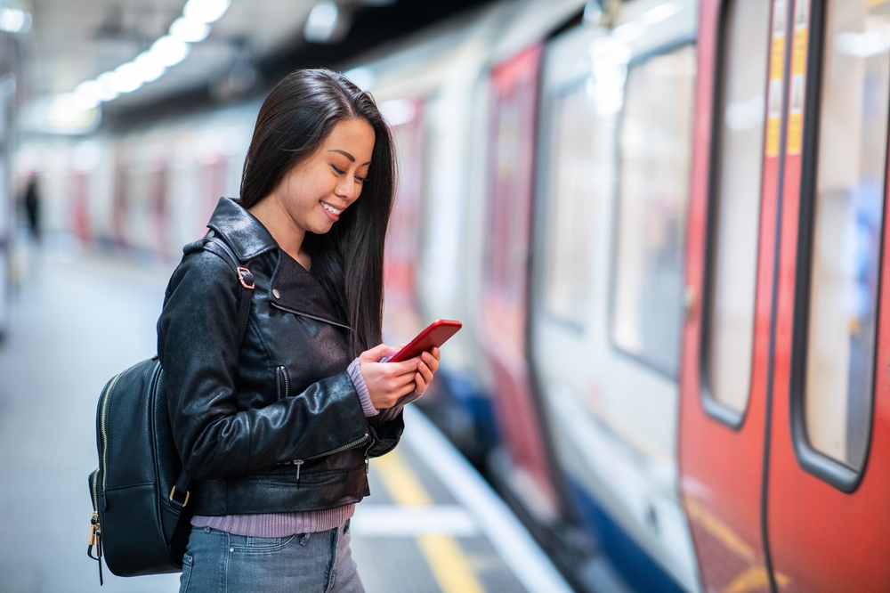 A young girl stands on the edge of the Tube platform, waiting for public transport with her leather jacket over her purple sweater and her backpack.