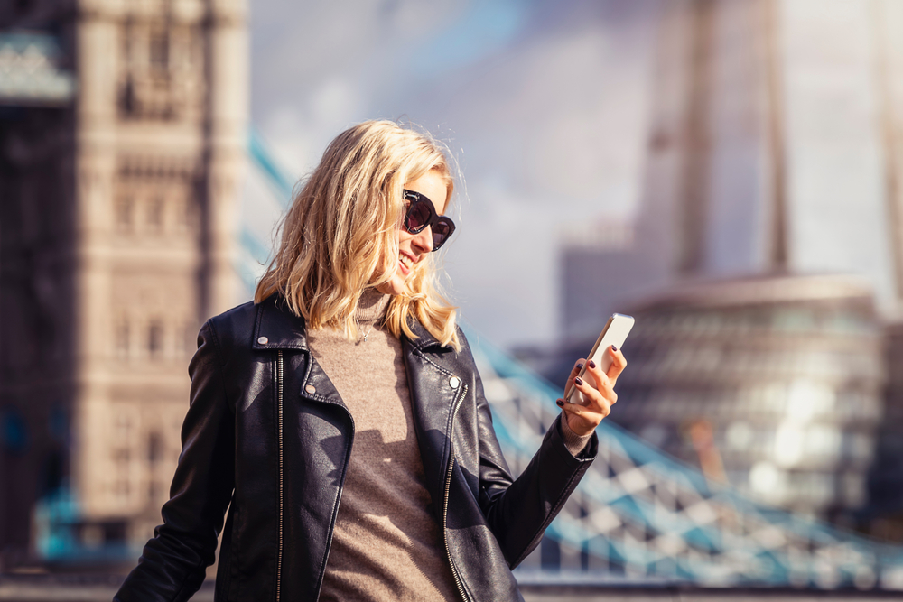 A young woman knows what to wear in London in September in her turtle neck, glasses, and leather jacket as she stands by tower bridge on her phone.