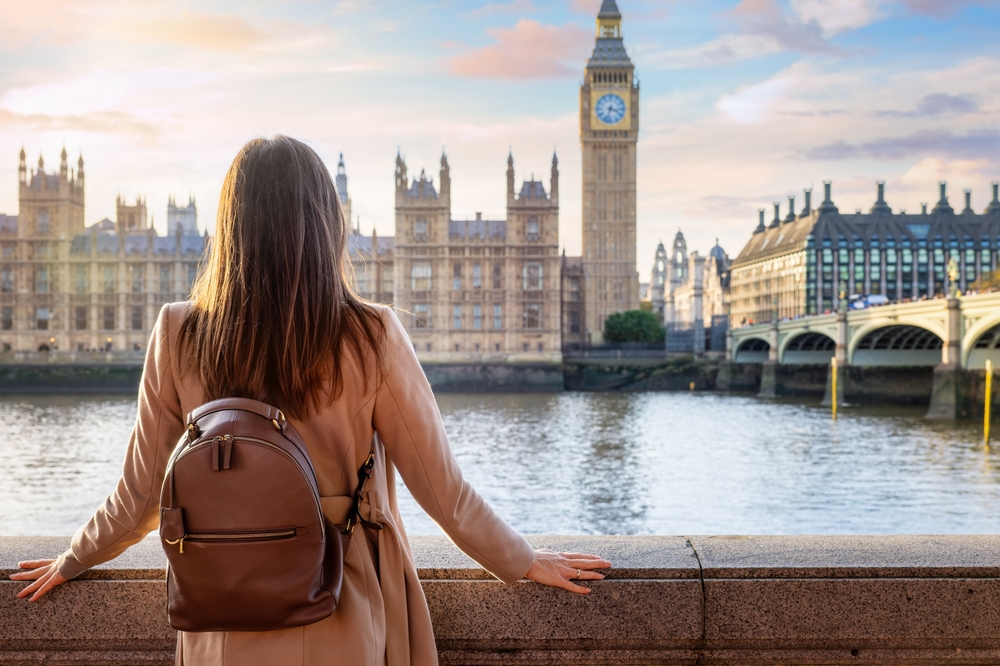 A woman stands across from Big Ben and looks across the river while wearing a suede jacket and a leather backpack.