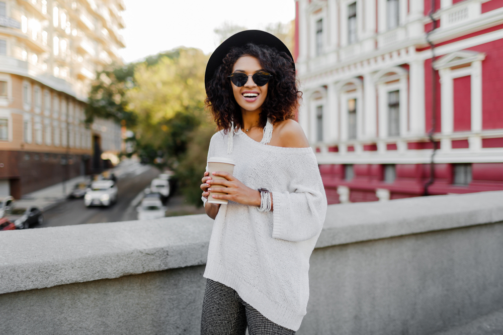 A young woman in an off-the-shoulder poncho holds a cup of coffee on a bridge in London.
