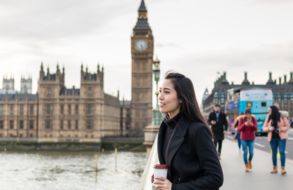 A young woman smiles as she leans over a bridge, looking at Westminster, knowing what to wear in London in fall in her black turtle neck and black peacoat while clutching her coffee.