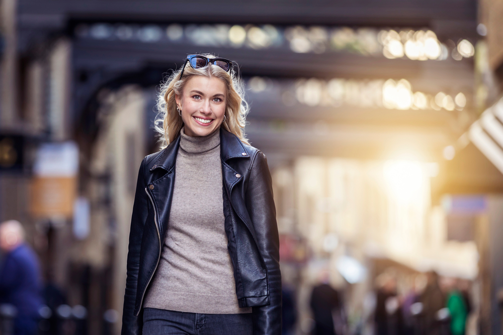 A woman smiles as she walks down the street of a market in London, her wool turtleneck under a leather jacket.