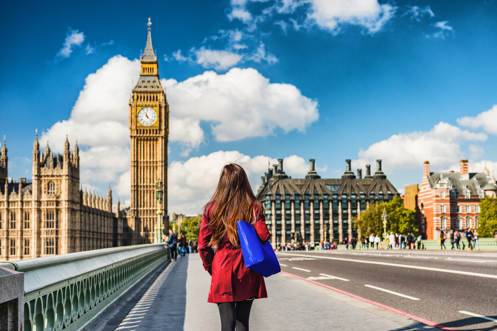 A woman knows what to wear in London in fall as she walks toward Westminster Abby in black leggings, a red trench coat and a blue tote on her arm.