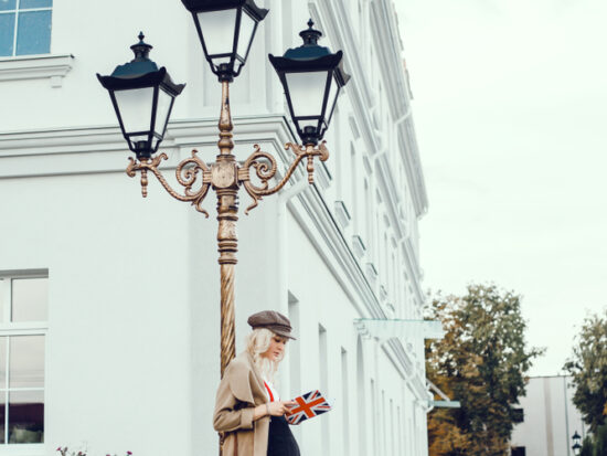 A woman leans against a lightpost in London, knowing what to wear in London in fall, in her pants , trench coat, and beret.