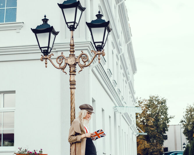 A woman leans against a lightpost in London, knowing what to wear in London in fall, in her pants , trench coat, and beret.