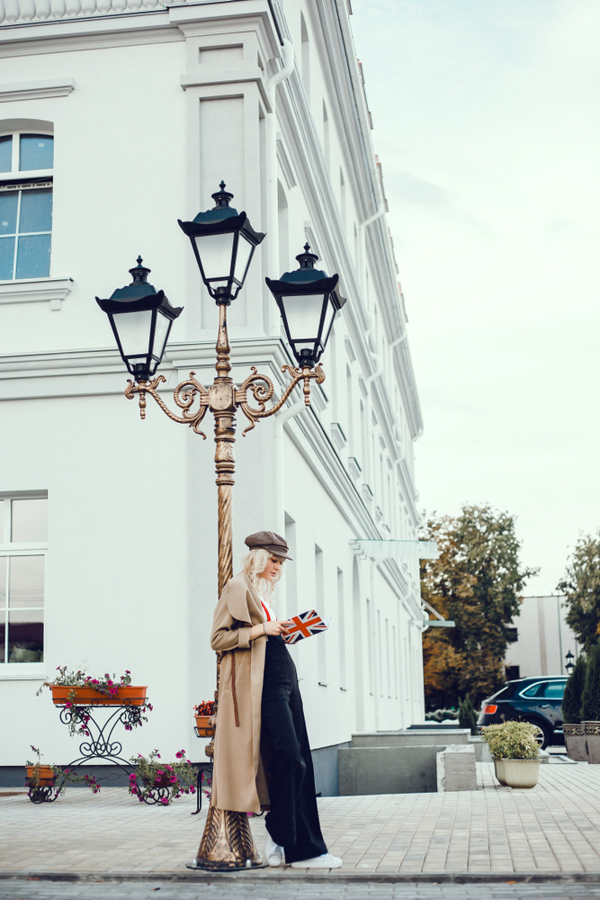 A young woman knows what to wear in London in fall as she wears a beige trench coat, a matching hat, a white short and black trousers with red suspenders and white sneakers as she leans against a random light post in the city.