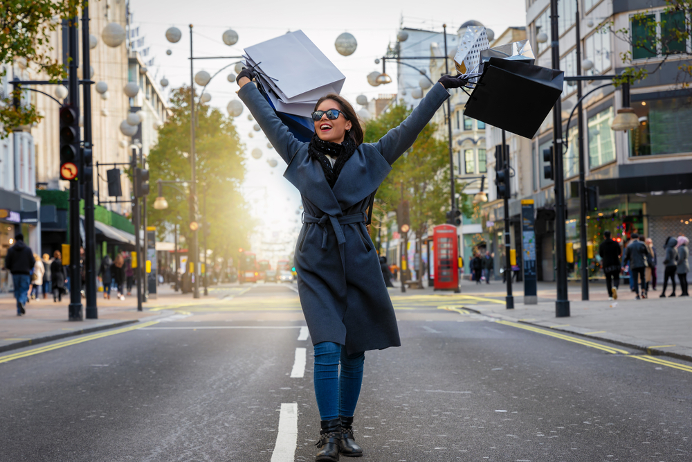 A woman walks down the street in London, shopping bags thrown in the air in excitement as she knows what to wear in London in fall, which includes a wool trench coat, scarf, jeans and even sunglasses.