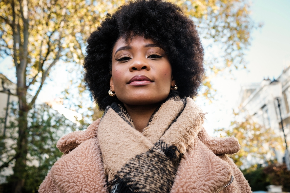 A woman looks directly at the camera with gold earrings, a fluffy coat, and a warm blanket scarf around her neck as the trees behind her shift colors: she knew what to wear in London in fall!
