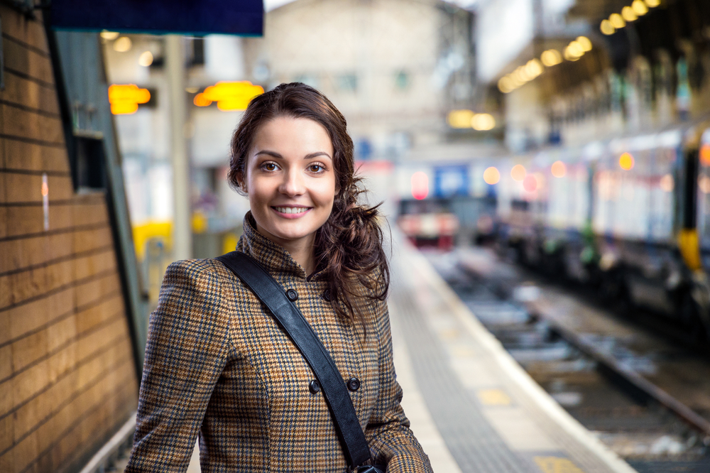 A young woman with curly brown hair smiles at the camera while awaiting public transport, wearing yellow tweed jacket and a crossbody bag.