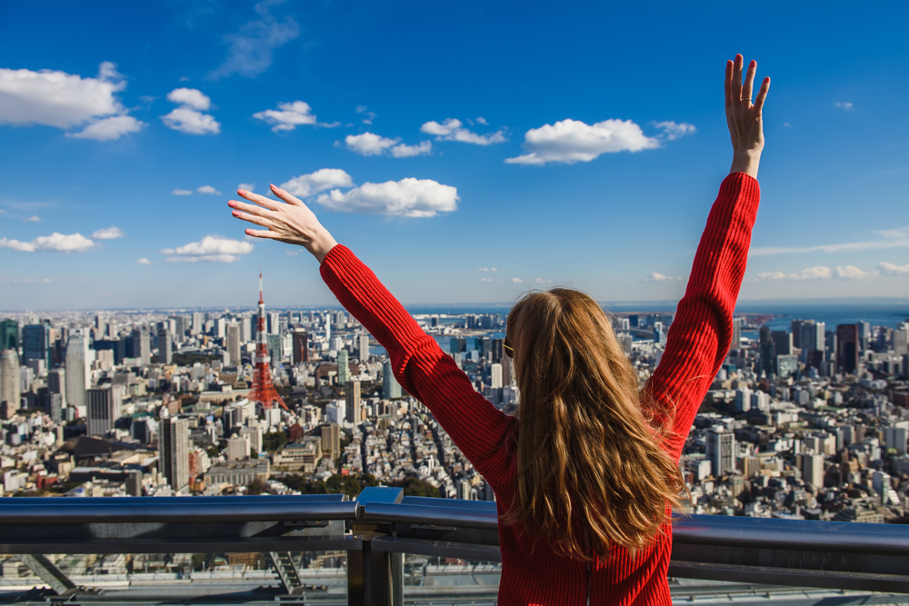 From a high viewpoint of Tokyo a woman in a red sweater puts her hands in the air as she looks down at the cityscape while wearing sunglasses.