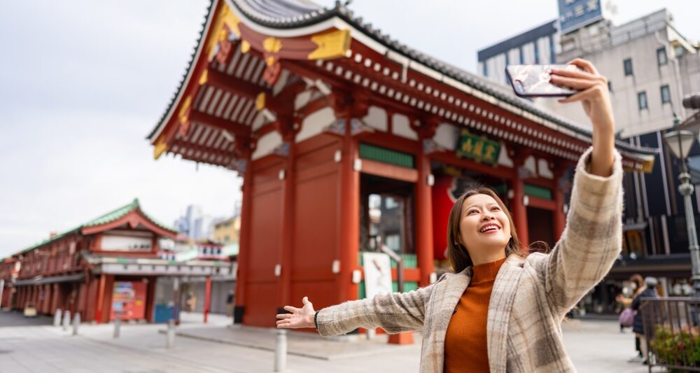 A woman in a plaid wool jacket over a turtleneck knows what to wear in Tokyo as she takes a selfie during the fall season at a famous temple in Japan.