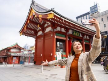 A woman in a plaid wool jacket over a turtleneck knows what to wear in Tokyo as she takes a selfie during the fall season at a famous temple in Japan.