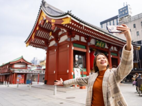 A woman in a plaid wool jacket over a turtleneck knows what to wear in Tokyo as she takes a selfie during the fall season at a famous temple in Japan.