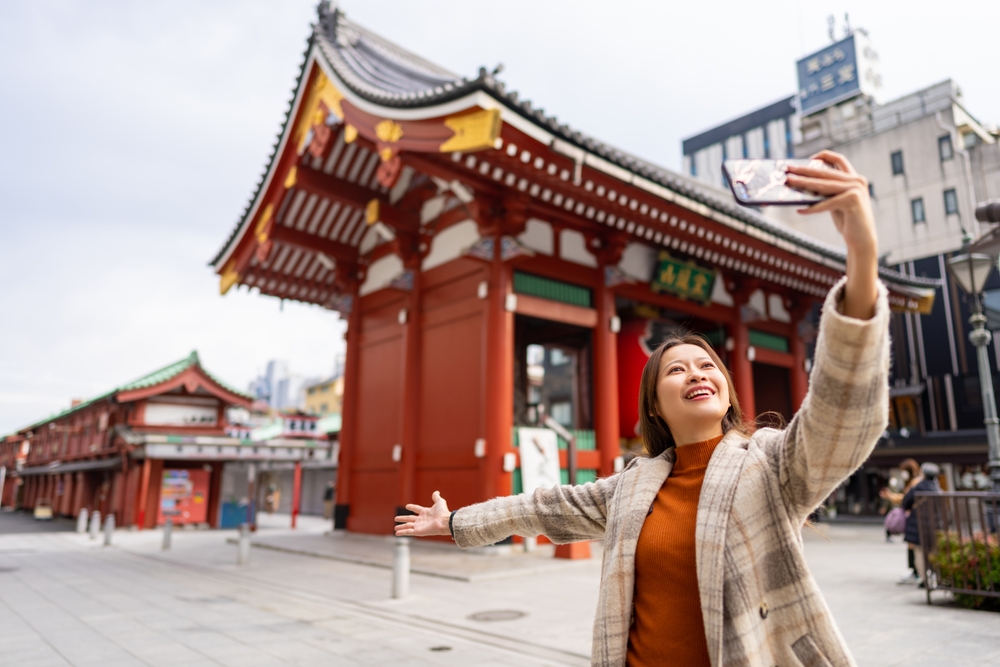 A woman knows what to wear in Tokyo in winter in her plaid, heavy outer wear, and her turtleneck as she holds up her phone for selfies at a temple.