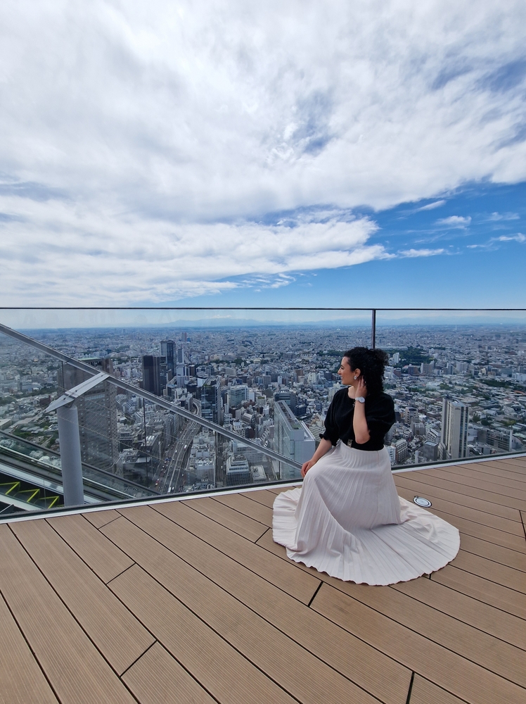 From a skyway viewpoint, a woman sits on an edge and overlooks the city, knowing what to wear in Tokyo in her pleated skirt and black blouse.