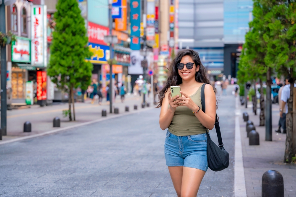 A woman with dark hair enjoys the summer weather in Tokyo as she walks down the street in a green tank top, green shorts, and a crossbody bag over her shoulder.