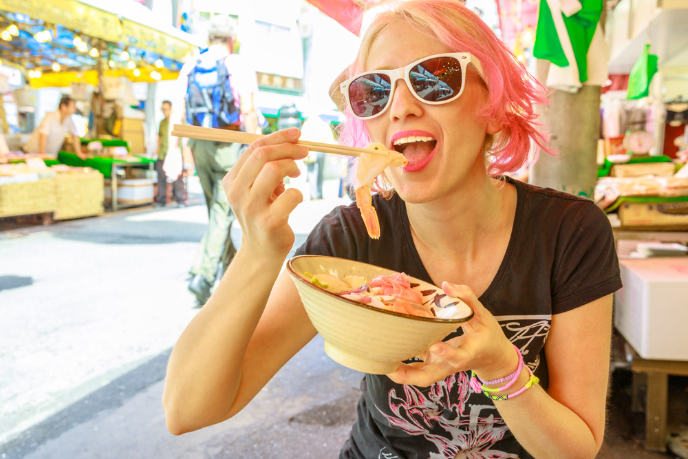 A woman with pink hair holds a bowl of food close to her mouth and in one hand has chopsticks, but she really knows what to wear in Tokyo to keep it casual and comfortable at this market in her graphic tee and sunglasses.