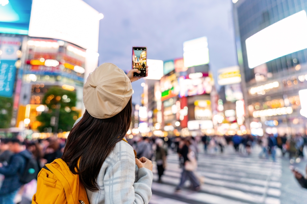 A woman in a plaid wool coat, yellow backpack and neutral, leather beret holds up her phone to take a photo in the main city hub.
