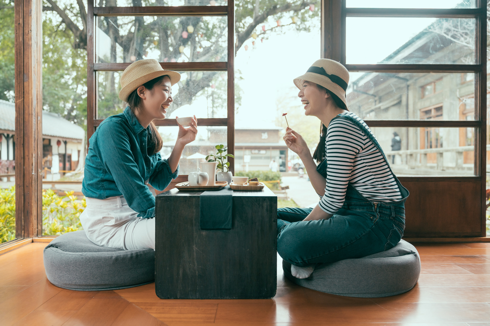 Two friends sit across from each other, eating and drinking tea, both knowing what to wear in Tokyo in a casual sense with their trousers, buttons downs, stripped tees and hats.