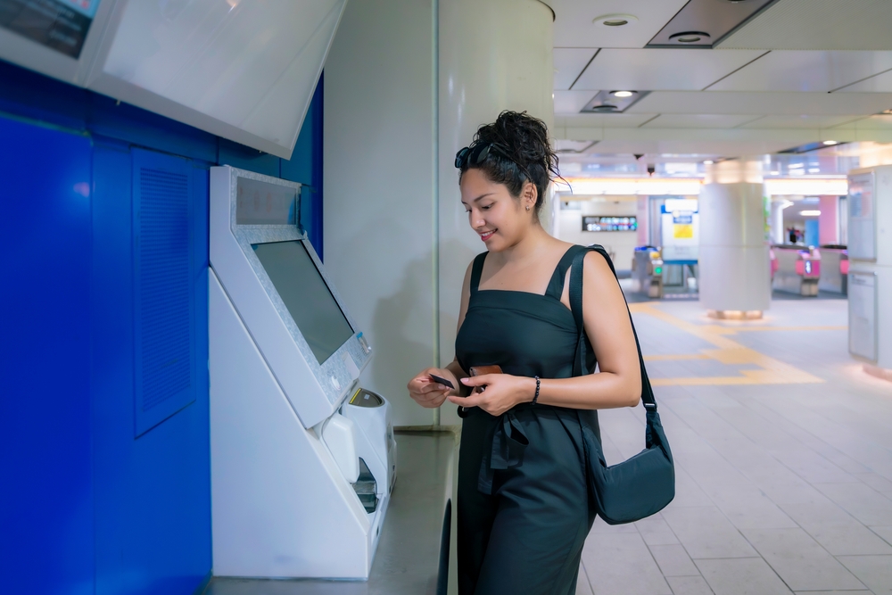 A woman in a black jumper stands a a kisok, buying public transportation tickets.
