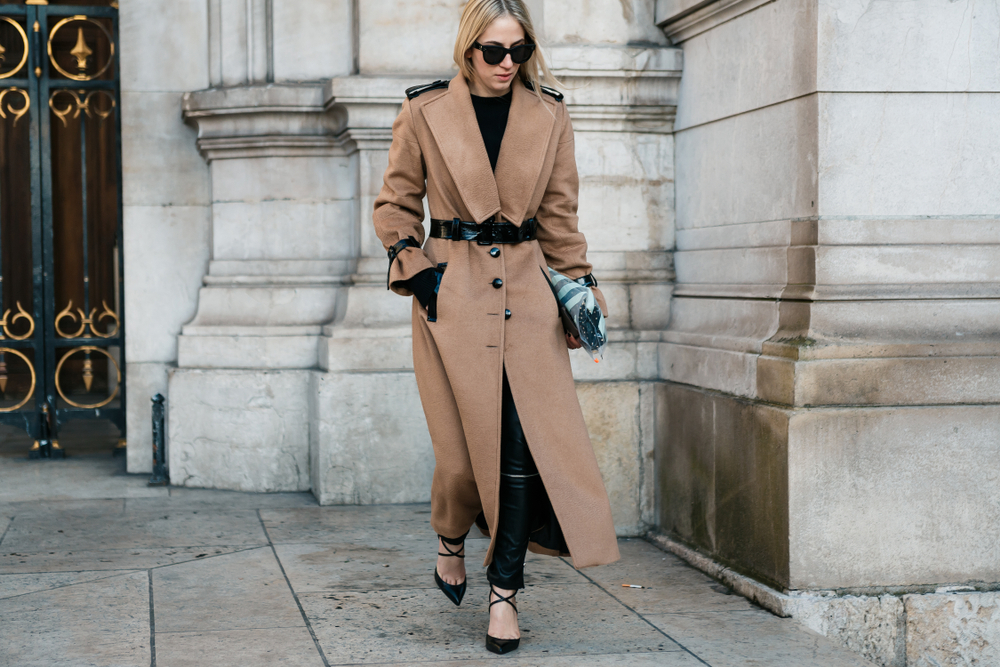 A woman stands in the streets wearing a trench coat, prepared for the fall, knowing what to wear to Paris in September.