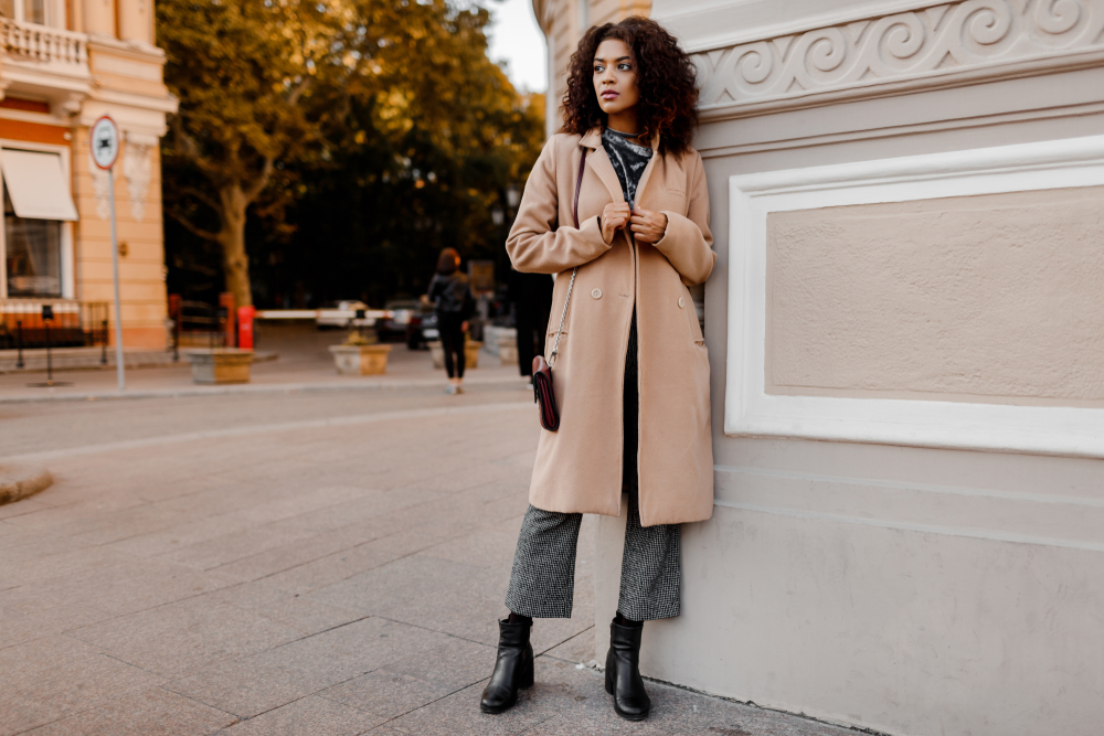 A woman in a long beige trench coat leans against a Paris monument, knowing what to wear to Paris in September with her black pants and ankle boots.