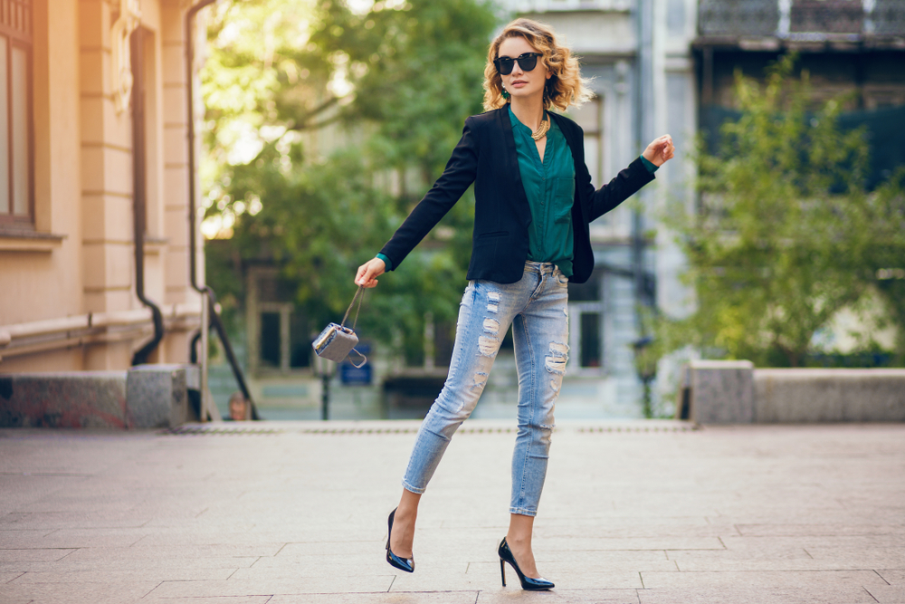 A woman dances in the streets of Paris since she knows what to wear to Paris in September in her jeans, silk blouse, heels, blazer and clutch.