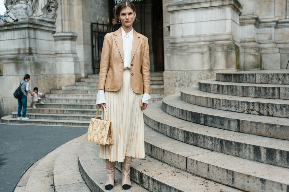 A woman in a blazer, button up and pleated maxi skit stands in Paris on old, historic steps.