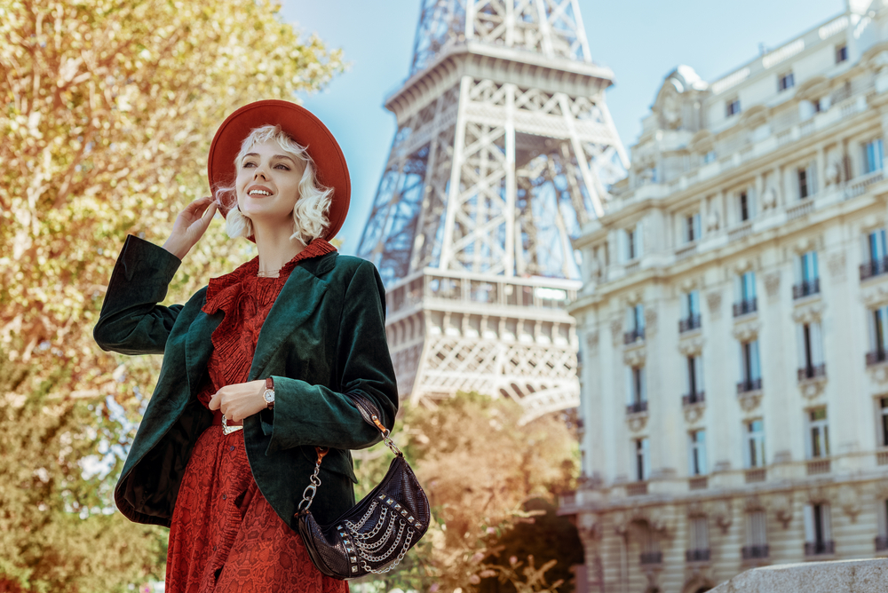 A woman in a green blazer, red dress and matching red hat smiles next to the Eiffel tower.